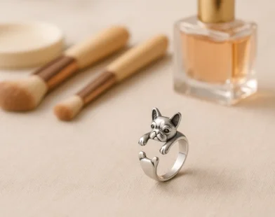 Detailed silver French Bulldog ring with wraparound design, styled beside makeup brushes and perfume for a chic look.