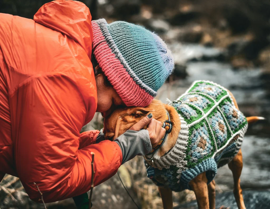 Person in orange jacket cuddling a small dog in a green and blue crochet sweater outdoors.